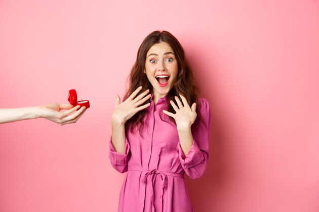 婚礼惊讶女友的画像兴奋的尖叫 手拿订婚戒指 接受求婚 看着相机惊讶 站在粉红色的墙上新娘女人年轻