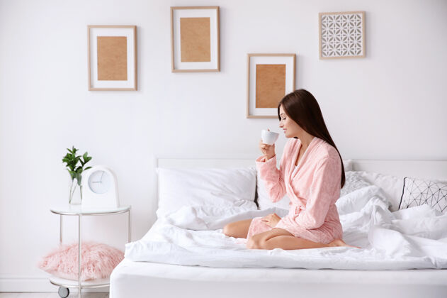 晨衣年轻女子坐在家里的床上喝咖啡觉醒懒惰醒