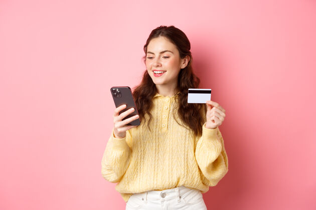 情绪科技和网上购物年轻漂亮的女士用信用卡在线支付 看着智能手机 微笑着 站在粉红色的墙上聊天应用程序候选人