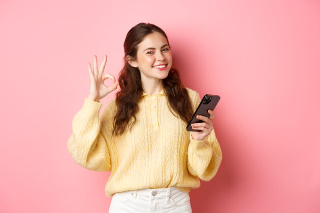 聊天科技和网上购物年轻的女模特展示好的标志和使用手机应用程序 称赞好的商店 说是的 站在粉红色的墙上互联网手势科技