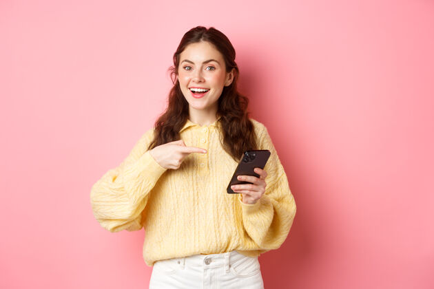 旁白年轻漂亮的女人拿着智能手机 指着屏幕 推销 谈论她的社交媒体页面 站在粉色的墙上欢呼在线手机