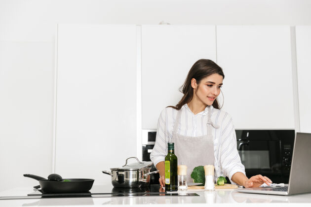 食物漂亮的年轻女子在厨房的锅里煮健康的晚餐 看着笔记本电脑站着白种人女人