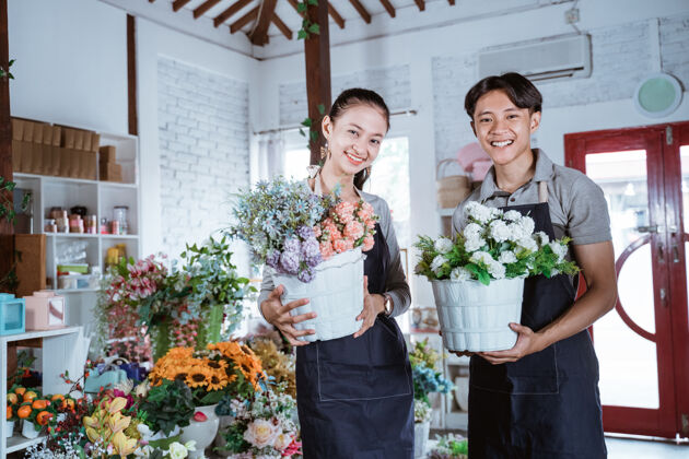 商业幸福的年轻夫妇穿着围裙拿着桶花微笑着看着相机在花店工作企业家花店团队合作