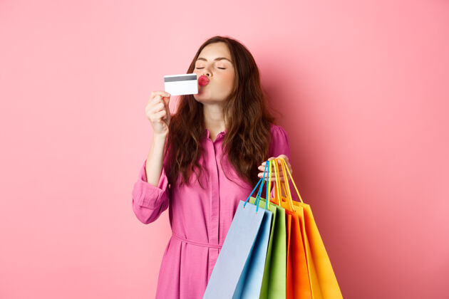 信用卡兴奋快乐的购物狂的画像 拿着购物袋和亲吻塑料信用卡的女人 面带惊奇的微笑 站在粉红色的墙上女孩候选人黑发