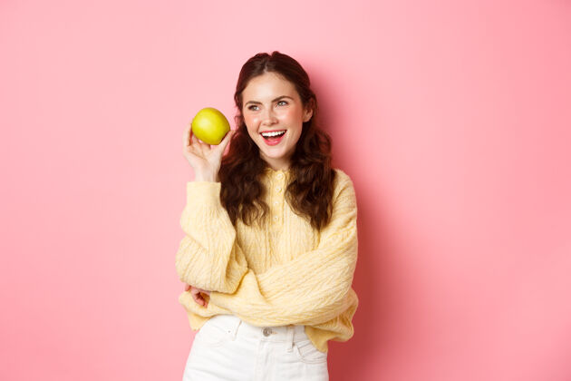 快乐健康和生活理念开朗微笑的女人 展现她洁白完美的笑容和绿色的苹果 吃着健康的水果 站在粉色的墙上成人女孩女人