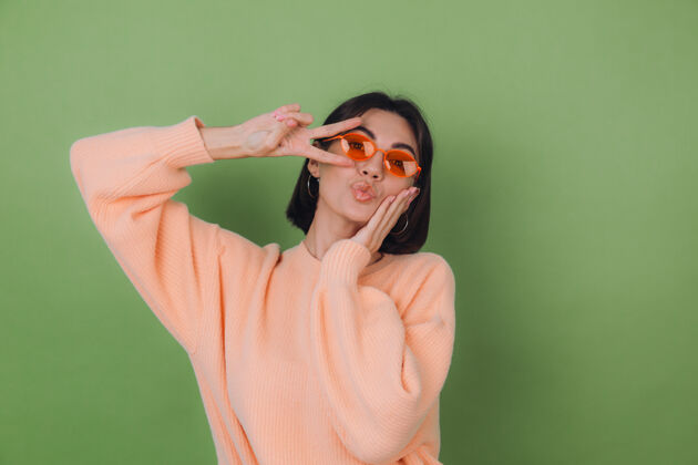 休闲年轻女子穿着休闲的桃色毛衣 戴着橙色眼镜 隔着绿橄榄色的墙壁滑稽地做着胜利手势复制空间欢乐成功脸