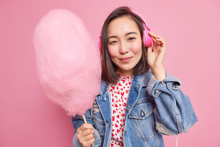 成人人们和生活方式的概念漂亮的亚洲少女通过耳机听音乐拿着美味的棉花糖穿着时尚的牛仔夹克享受自由时间隔离在粉红色的墙上触摸感觉音乐
