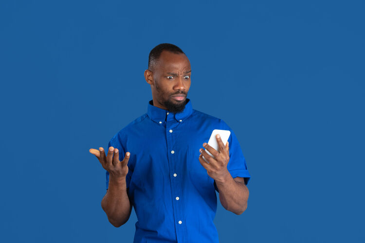 表情想知道如何使用电话隔离在蓝色墙上的非洲裔美国年轻人的单色肖像非洲男人手势