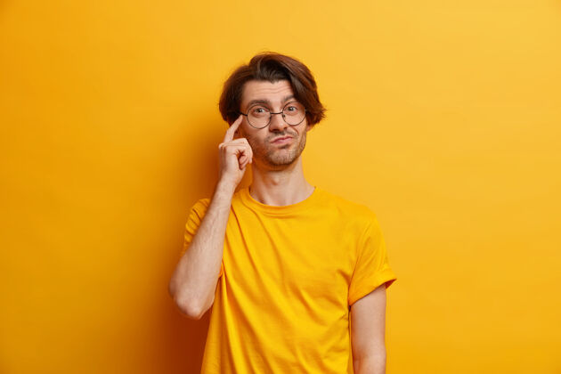 休闲体贴的年轻欧洲人的照片把手指放在太阳穴上想象着有人戴着圆眼镜和休闲t恤隔离在黄色的墙上做重要的决定时尚表情男人
