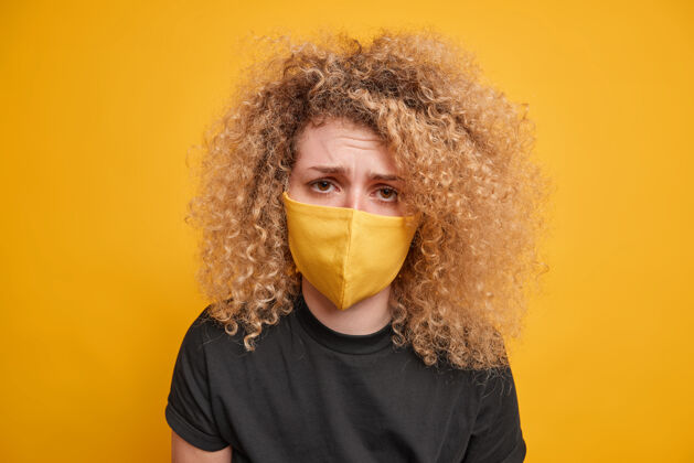 面具一个卷发女人的特写镜头看起来很悲伤戴着防护面具厌倦了禁闭限制穿着黑色t恤隔离在黄色的墙上冠状病毒大流行人类姿势预防