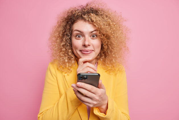 微笑美丽的卷发年轻女子手持手机在线阅读精彩新闻看起来很高兴使用免费互联网穿着黄色夹克隔离在粉红色的墙上科技概念短信脸短信