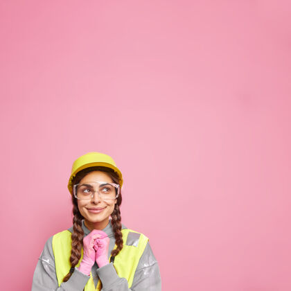 保护女建筑工助理双手合十 沉思地看着上面 想着新建筑的施工 戴着防护头盔 戴着安全眼镜 制服隔着粉红色的墙安全帽职业穿