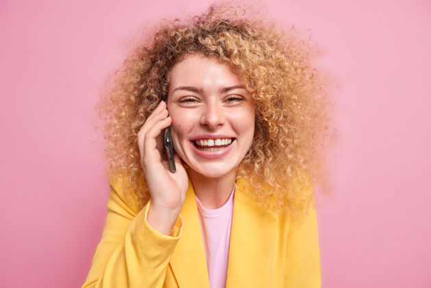 教育积极的卷发女模特有着快乐的表情享受愉快的电话交谈微笑着牙关露出牙齿穿着黄色夹克隔离在粉红色的墙上交谈时咯咯笑设备女人说话
