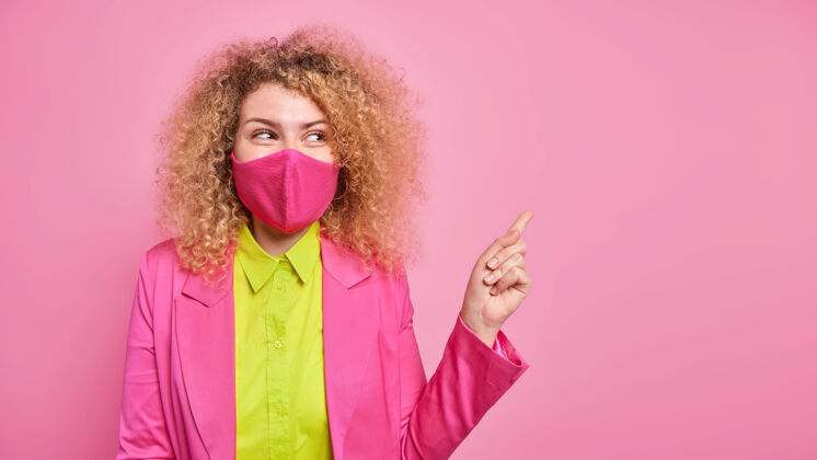 隔离漂亮开朗的卷发女人戴着口罩的水平镜头在右上角显示出你的广告空间被隔离在粉红色的墙上预防措施女性面具姿势