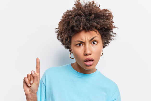 女性不高兴的卷发非裔美国妇女头像困惑地表示 食指以上显示一些惊人的穿着卡苏拉蓝色t恤隔离在白色的墙壁情感非洲黑发