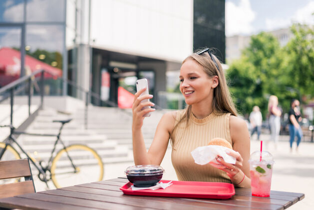 食物美丽的年轻女子拿着手机在户外咖啡馆吃着美味的汉堡营养饲料社交媒体