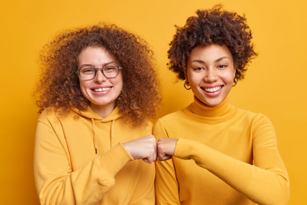 拳头两个快乐的不同的女人做拳头碰撞示范协议有友好的关系微笑愉快地站在彼此旁边隔着黄墙团队合作的肢体语言概念家庭成功欢呼