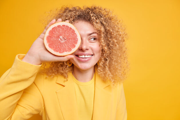 空有机饮食概念高兴的卷发女人用葡萄柚遮住眼睛半笑着牙关要做新鲜果汁或冰沙看着旁边的姿势靠着黄色的墙壁文本空间区域积极盖成人