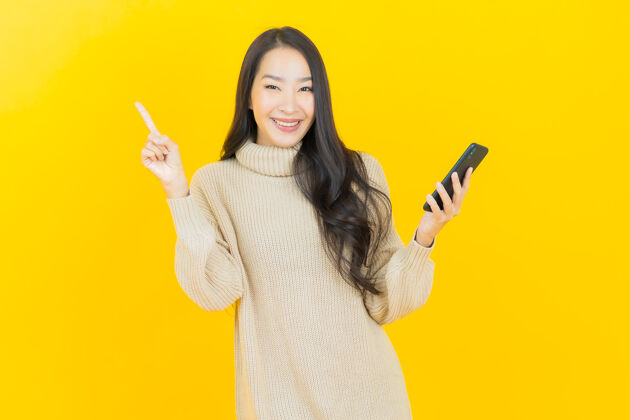 漂亮画像美丽的亚洲年轻女子微笑着在黄色的墙上智能手机手指电话商务