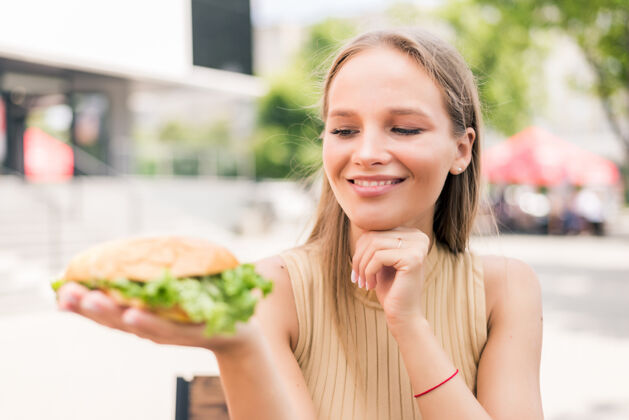 美丽年轻漂亮的女人拿着汉堡包坐在户外饥饿面包城市