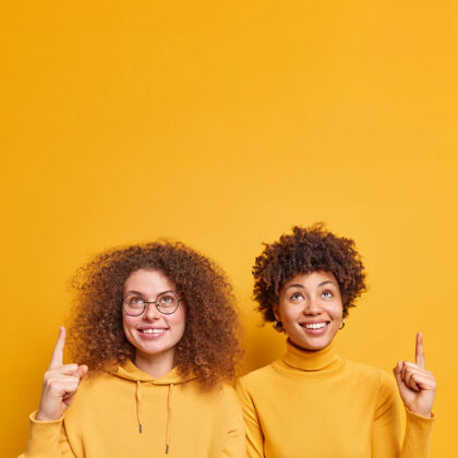 女士快乐的年轻多样化的女性镜头点在复制空间的上方 礼品或产品隔着黄色的墙壁相互独立地立着人们宣传和广告理念肖像女人休闲