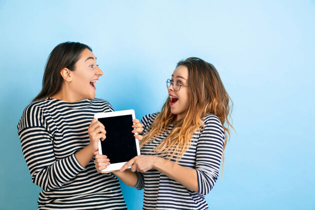 人类手持空白屏幕的平板电脑在渐变蓝色工作室背景上孤立的年轻情感女性人类情感的概念 面部表情 友谊 广告穿着休闲服的美丽模特女孩表情在一起