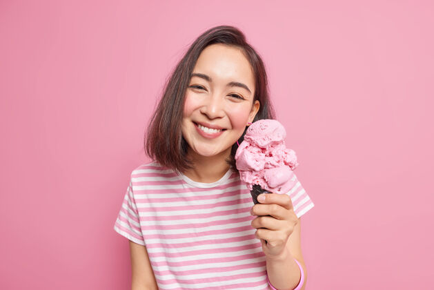 条纹可爱的深色头发的亚洲女人喜欢在炎热的夏天吃美味的草莓冰淇淋 心情乐观 表情愉悦 穿着随意 在室内摆出最喜欢的冷冻甜点人甜点拿着