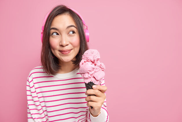 女性长着一头黑发的可爱的亚洲年轻女子拿着大大的草莓冰淇淋 若有所思地移开视线 戴着立体声耳机 隔着粉色的墙壁空间衣服模特立体声