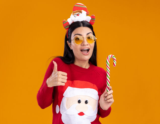 头带令人印象深刻的年轻白人女孩戴着圣诞老人的头带和毛衣 戴着眼镜 手持传统的圣诞糖果手杖 竖起大拇指 孤立地站在橙色的墙上 留着复制空间年轻女孩白种人