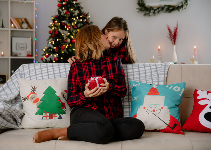 亲吻高兴的母亲拿着礼盒 亲吻坐在沙发上的女儿 在家里享受圣诞节时光抱着女儿时间