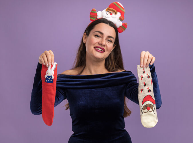 年轻微笑着的年轻漂亮的女孩穿着蓝色的裙子 戴着圣诞发箍 手里拿着圣诞袜 孤立地站在紫色的墙上圣诞节微笑女孩