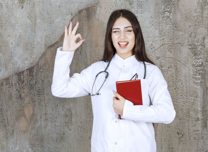 护理面带微笑的女医生姿势很好 拿着书站在灰色的墙上医疗保健女性女性