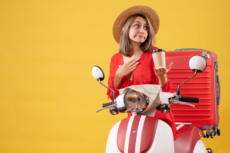 行李正面图身穿红色连衣裙的年轻女士拿着咖啡杯 手放在她的下巴上 靠近轻便摩托车下巴手旅游