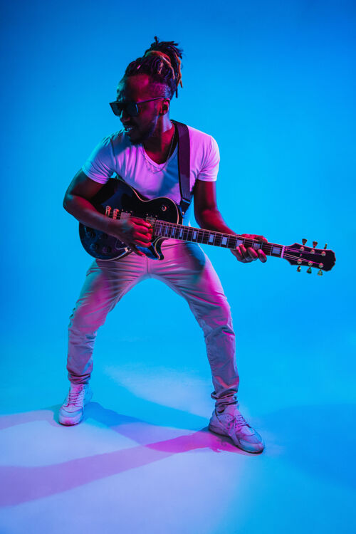 娱乐年轻的非洲裔美国音乐家在霓虹灯下像摇滚明星一样在蓝色的工作室背景上弹吉他音乐的概念 爱好快乐的家伙即兴创作复古的彩色肖像月灯光旋律