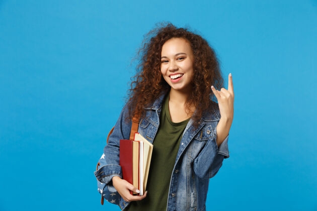 微笑穿着牛仔服 背着书包的年轻女学生把书孤立地放在蓝色的墙上知识摇滚快乐