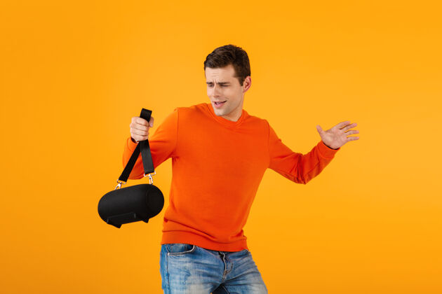 声音时尚微笑的年轻人穿着橙色毛衣手持无线扬声器快乐地听音乐玩得开心情感搞笑科技