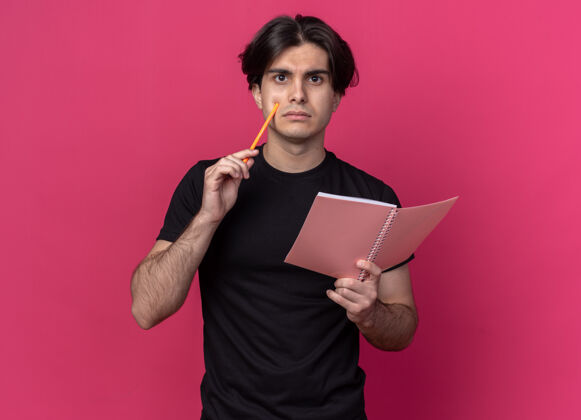 市民想着年轻帅哥穿着黑色t恤拿着笔记本用铅笔隔离在粉红色的墙上人表情姿势