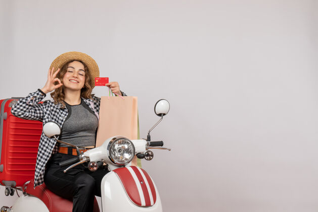 信用卡正面图：骑着轻便摩托车的年轻女子拿着卡片做着“ok”标志坐着灰色成人