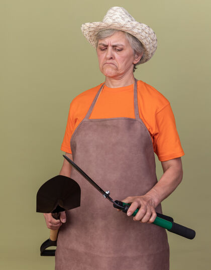 绿色自信的上了年纪的女园丁戴着园艺帽拿着铲子看着园艺剪刀举行穿剪刀