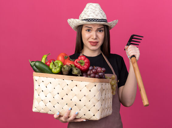 漂亮焦虑美丽的白人女园丁戴着园艺帽拿着菜篮和耙子举行耙帽子