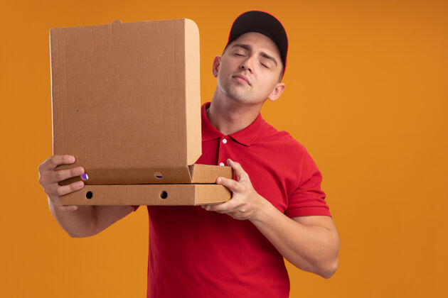 开喜欢闭着眼睛的年轻送货员穿着制服 戴着帽子 嗅着隔离在橙色墙上的披萨盒衣服眼睛人