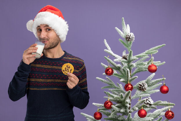 男人戴着圣诞帽的年轻白种人高兴地站在装饰好的圣诞树旁 手里拿着一杯牛奶和一块饼干 喝着隔离在紫色墙上的牛奶抱着帽子玻璃杯