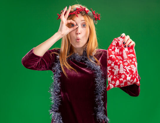 女孩惊讶的年轻漂亮的女孩穿着红色的衣服 脖子上戴着花环和花环 手里拿着圣诞包 在绿色的墙上显示出孤立的表情和姿态脖子显示穿