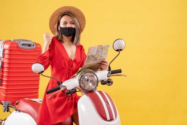 轻便摩托车正面图：戴着黑色面具的年轻女士手持地图 在轻便摩托车旁表达她的幸福持有摩托车游客