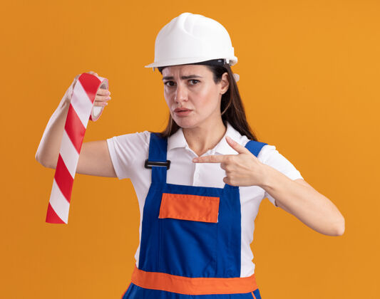 年轻一个穿着制服的严格的年轻的建筑女工人指着隔离在橙色墙上的管道胶带建设者感觉制服