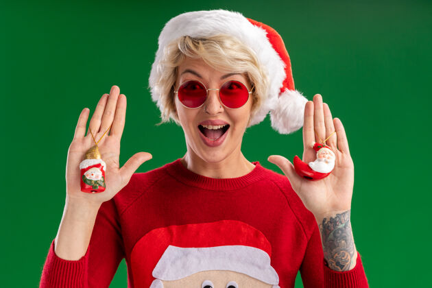 秀令人印象深刻的年轻金发女人戴着圣诞帽和圣诞老人圣诞毛衣与眼镜看雪人和圣诞老人圣诞饰品孤立在绿色的墙壁眼镜毛衣雪人