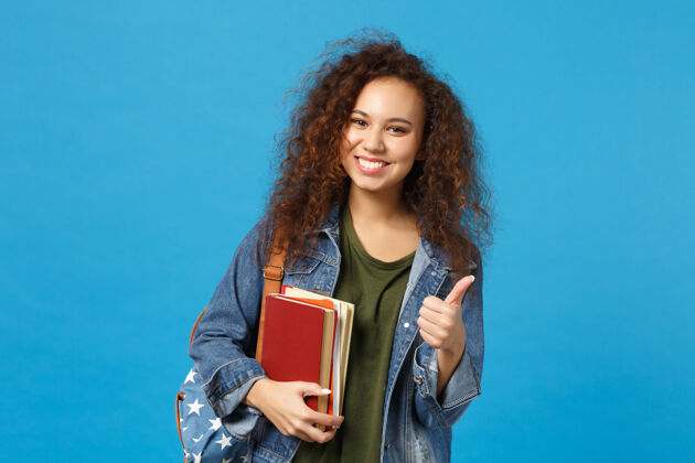 背包穿着牛仔服 背着书包的年轻女学生把书孤立地放在蓝色的墙上教学青少年背