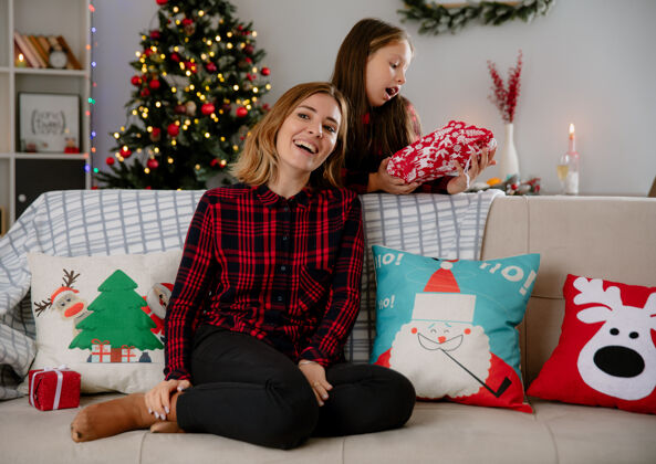 包装微笑的母亲看着坐在沙发上的一旁 兴奋的女儿拿着礼包 在家里享受圣诞节时光侧身享受房子
