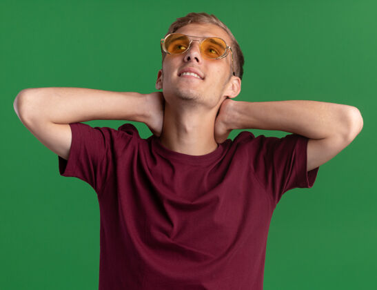 眼镜微笑着抬头看年轻帅哥穿着红衬衫和眼镜把手放在脖子上隔离在绿色的墙上穿着感觉衬衫