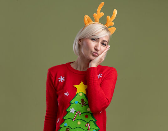 脸自信的中年金发女人 戴着圣诞驯鹿鹿角头带和圣诞毛衣 看起来手贴在脸上 做着亲吻的手势 在橄榄绿的墙上 留着复制空间毛衣金发中年